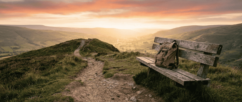 Sentier de crête qui continue vers une vallée lumineuse, banc et sac à dos au bord du chemin